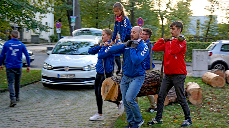 Vier Menschen tragen einen Stamm mit Bändern und Stangen. Ein Mädchen sitzt auf dem Stamm.