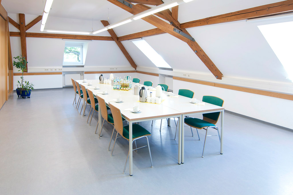 Seminarraum mit Holzbalken, Dachschrägen, Tische als geschlossener Block, Kaffee-/Teegedeck und Kaltgetränke auf den Tischen, Blick von der Tür