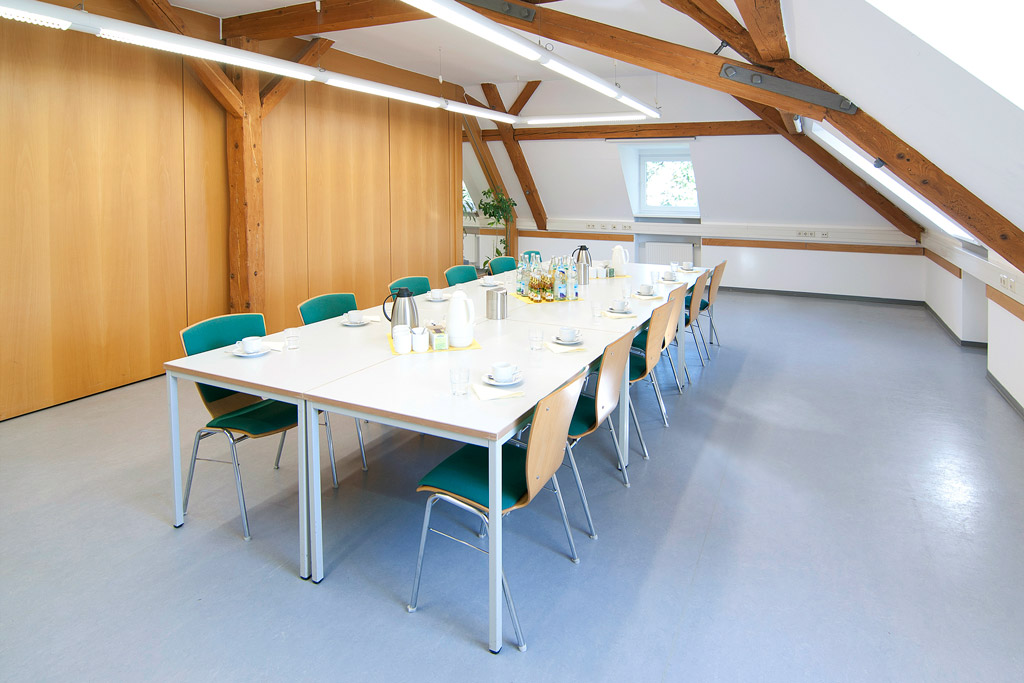 Seminarraum mit Holzbalken und Sicht auf Trennwand, Dachschrägen, Tische als geschlossener Block, Kaffee-/Teegedeck und Kaltgetränke auf den Tischen, Blick von der Fensterseite