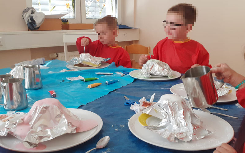 Kinder sitzen an einem Tisch. Jedes hat einen Teller vor sich mit einem Versuchsaufbau, um einen Vulkan zu simulieren