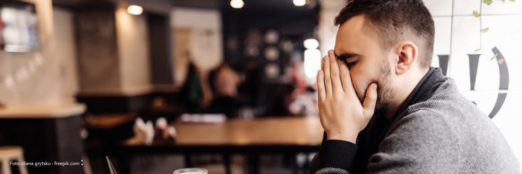 Mann sitzt in einem Kaffee, beugt das Gesicht in die Hände. Er wirkt erschöpft.